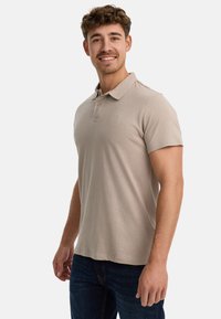 Beige Poloshirt aus weichem Baumwollmaterial, mit klassischem Kragen, zwei Knöpfen und einem dezenten Logo auf der Brust. Kombiniert mit dunklen Jeans.