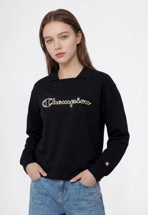 Ung kvinde med lysebrunt hår iført en sort Champion sweatshirt og blå jeans, hænderne i lommerne, mod en ensartet hvid baggrund.