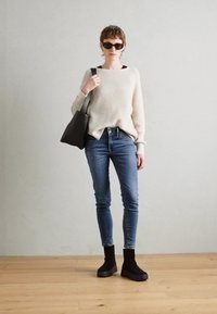 Pull côtelé crème, jean skinny bleu, bottines noires, et sac fourre-tout noir. Le modèle porte des lunettes de soleil sombres et se tient devant un fond neutre.