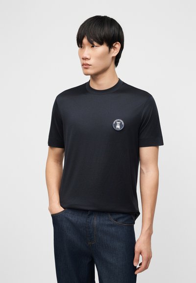 Άνδρας ντυμένος με μαύρο κοντομάνικο T-shirt που έχει μικρό στρογγυλό λογότυπο στο στήθος και σκούρο μπλε τζιν, που στέκεται μπροστά σε απλό ανοιχτόχρωμο φόντο.