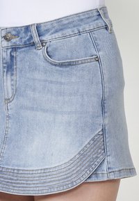 Denim mini krilo v svetlo modri barvi z ukrivljenim robom. Ima sprednje žepke, detajle šivanja in kovinsko gumbno zaklepanje.