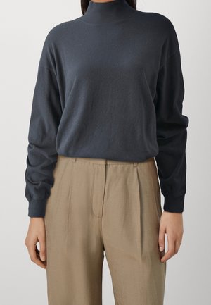 Personne portant un col roulé gris foncé à manches longues, rentré dans un pantalon beige plissé taille haute, debout les bras détendus.