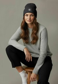 Black knit beanie with a logo, worn with a light gray sweater. Model has long, wavy hair and is seated on a wooden stool.
