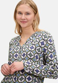 Hemdbluse mit geometrischem Muster in Blau, Grün und Weiß, einem V-Ausschnitt und geknöpften langen Ärmeln. Glatte Stofftextur.