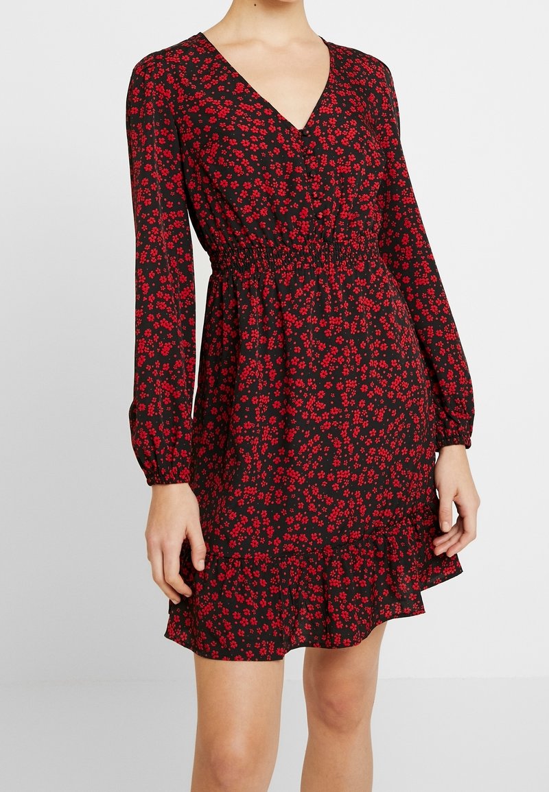 Vestido negro de manga larga, con escote en V, cintura fruncida y patrón floral rojo. El dobladillo con volantes añade textura y detalle.
