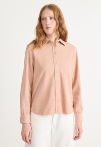 Long-sleeve, button-up shirt in light pink corduroy, featuring a classic collar and rounded hem, with visible stitching and soft texture.