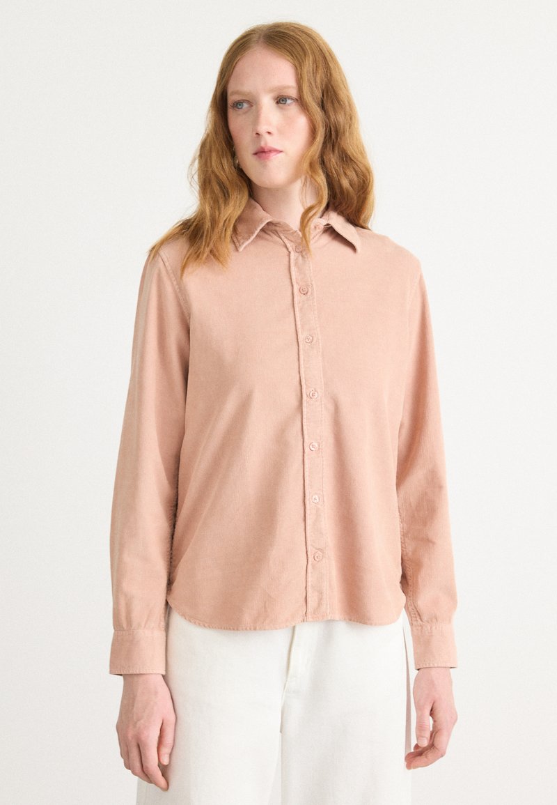 Long-sleeve, button-up shirt in light pink corduroy, featuring a classic collar and rounded hem, with visible stitching and soft texture.