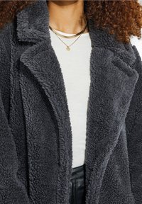 Donna che indossa un cappotto in pile testurizzato grigio scuro, una maglietta bianca, pantaloni neri e collane d'oro sovrapposte.