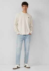 Lichtbeige lange mouw shirt met een geribbelde textuur, gecombineerd met lichtblauwe denim jeans en witte sneakers met zwarte accenten.