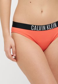 Calvin Klein bikinių trumpikės iš dryžuoto koralinio audinio. Juodos elastingos juostos su "CALVIN KLEIN" logotipu. Artimas šoninis ir priekinių vaizdo kampas.