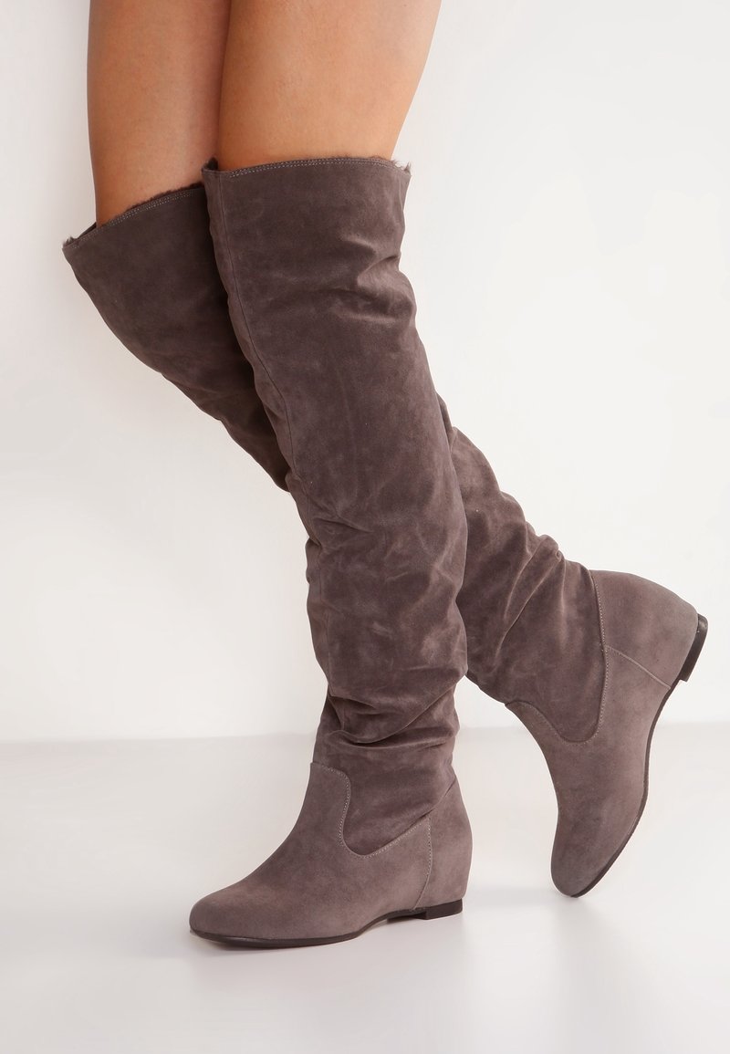 Femme portant des bottes compensées grises en daim montant jusqu'aux genoux, debout avec un pied légèrement levé sur un fond blanc.