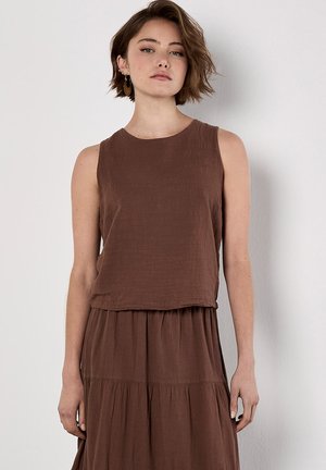 Model with short, tousled hair poses in a sleeveless brown dress against a neutral background, showcasing a relaxed yet stylish look.
