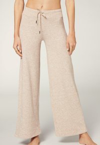 Kvinna som bär beige vida, dragsko loungebyxor stående barfota mot en ljus bakgrund.