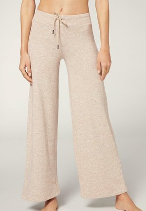 Femme portant un pantalon de lounge beige à jambes larges et à cordon de serrage, debout pieds nus devant un fond clair.