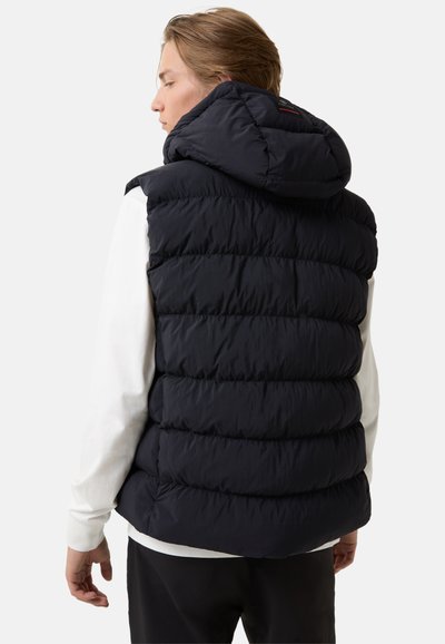 Námořnická pufovaná vesta s kapucí, která má prošívané části pro izolaci. Tkanina má lesklou texturu a je kombinována s bílou košilí s dlouhým rukávem.