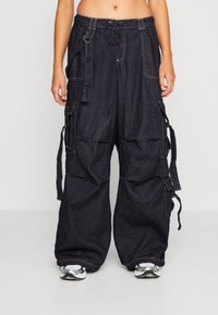 Dark denim cargo pants with a relaxed fit, featuring multiple side pockets, adjustable ties, and contrasting stitching details.