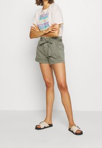 Shorts de talle alto color verde oliva con cinturón, dos bolsillos laterales, combinados con una camiseta gráfica clara y sandalias beige con un detalle de hebilla.