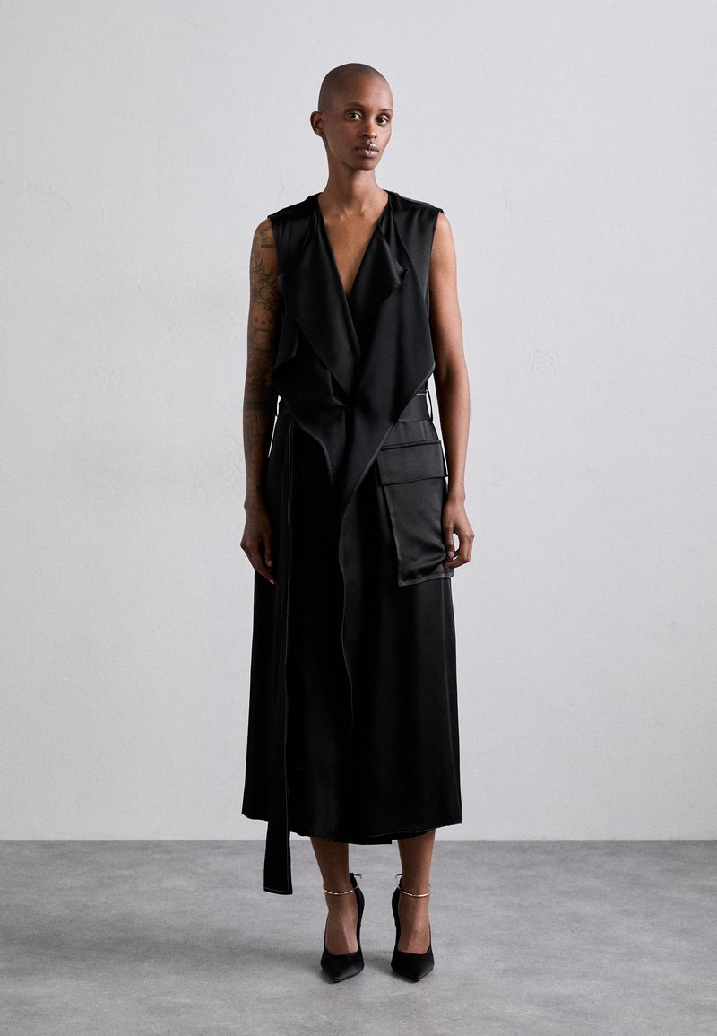 Black sleeveless dress with a draped front, featuring a pocket detail and a long tie. Smooth fabric, mid-calf length, worn with stiletto heels.