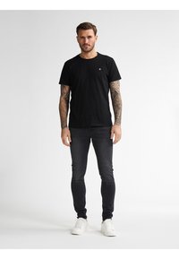 Camiseta de algodón negra de manga corta, con cuello redondo, que presenta un pequeño logotipo rojo en el pecho. Llevada con jeans ajustados oscuros y zapatillas blancas.