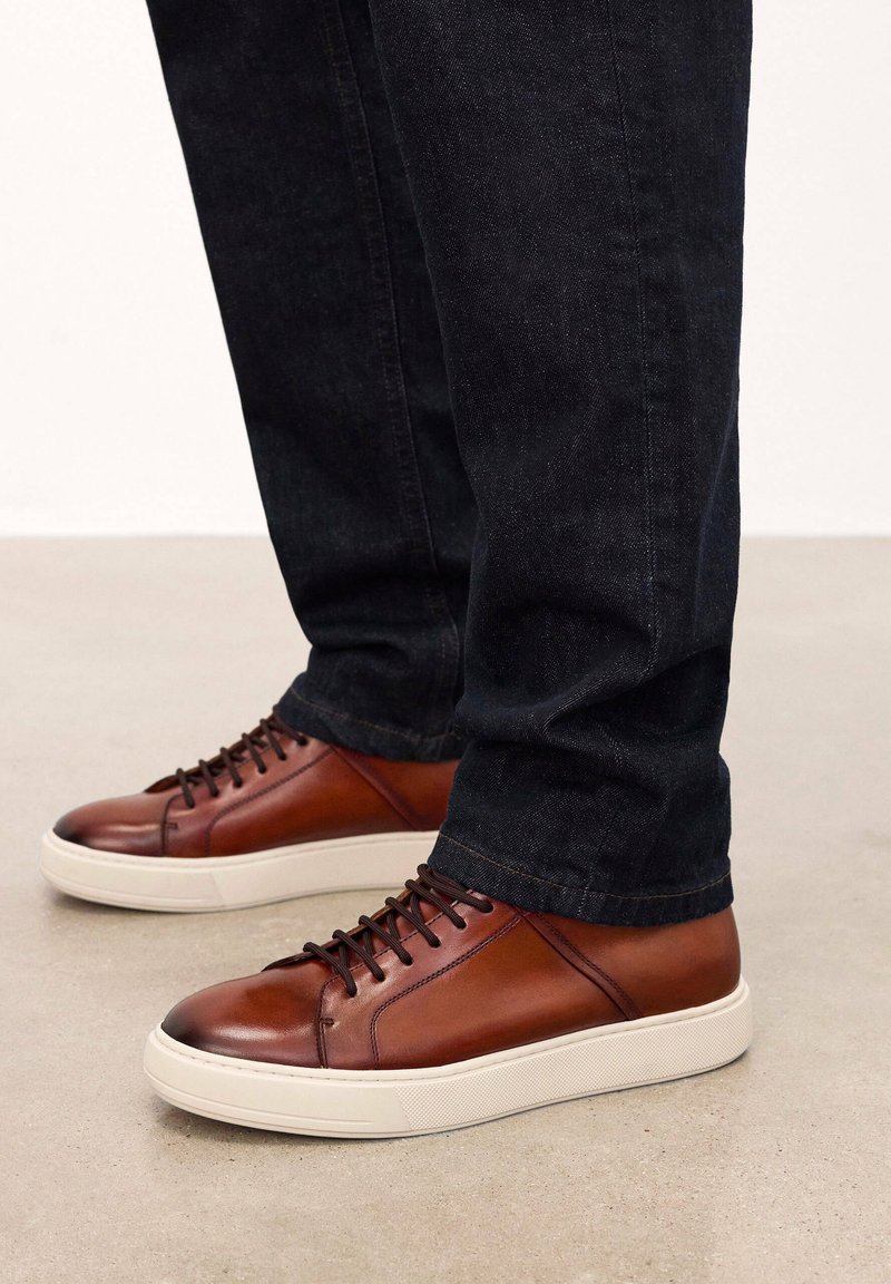 Zapatillas de cuero marrón con una textura suave, que presentan una suela de goma blanca y cordones marrones, combinadas con unos jeans de mezclilla oscura.