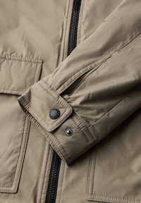 Manche de veste beige avec des boutons-pression réglables au poignet et une fermeture éclair noire verticale, montrant des coutures détaillées et la texture du tissu.