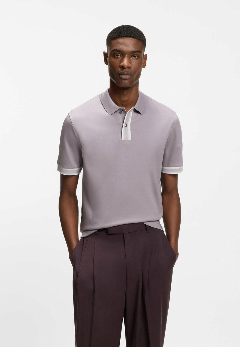 Homme portant un polo gris clair avec des bandes blanches sur les manches et le plastron, associé à un pantalon plissé violet foncé, les mains dans les poches.