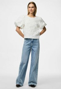 Top corto bianco testurizzato con maniche a palloncino abbinato a jeans azzurri a vita alta e a gamba larga. Scarpe nere completano l'outfit.