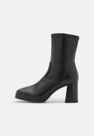 Bottine en cuir noir avec un bout carré arrondi, des coutures sur le panneau latéral et un talon bloc épais sur un fond blanc.