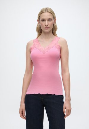 Vrouw met lang blond haar, gekleed in een mouwloos topje met roze kanten rand en donkerblauwe spijkerbroek, staand tegen een effen witte achtergrond.