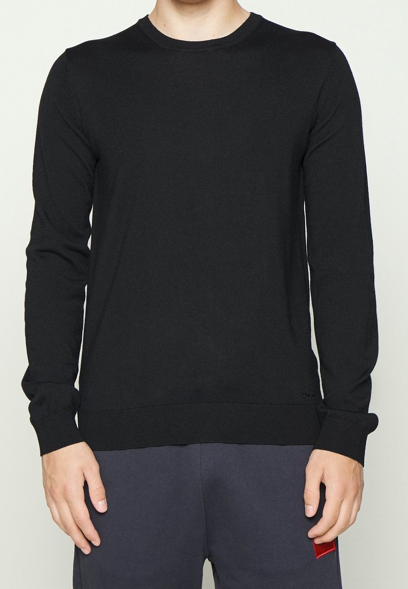 Homme portant un pull noir uni à manches longues et col rond, ainsi qu’un pantalon foncé, debout avec les bras détendus le long du corps sur un fond neutre.