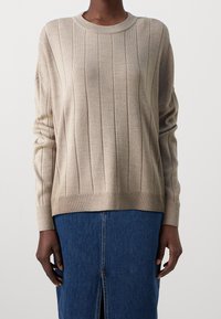 Beige ribbattuida neule, jossa on pyöreä kaula-aukko, pitkät hihat ja suora helma. Ominaisuuksiin kuuluvat pystysuorat raidat ja pehmeä rakenne. Yhdistetty farkkuhameeseen.