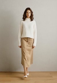 Pulover alb cu dungi din tricot, cu guler înalt, asortat cu o fustă deschisă la culoare, cu nasturi, și tocuri albe, stând pe un podea din lemn, pe fundal neutru.