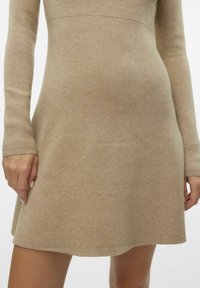 Abito lungo a maniche lunghe di maglia beige con corpetto aderente e gonna svasata. Tessuto morbido e design semplice, senza motivi o grafiche visibili.