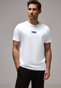 Uomo che indossa una maglietta bianca a maniche corte con un piccolo logo rettangolare nero sul petto, abbinata a pantaloni neri, su uno sfondo grigio neutro.