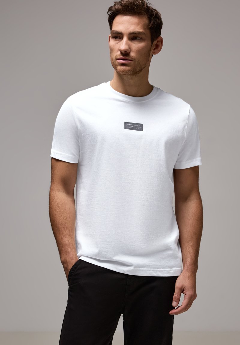 Uomo che indossa una maglietta bianca a maniche corte con un piccolo logo rettangolare nero sul petto, abbinata a pantaloni neri, su uno sfondo grigio neutro.