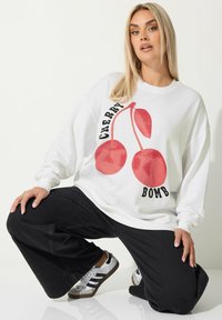 Vit sweatshirt med röd körsbärsgrafik och texten "Cherry Bomb". Modellen bär svarta vida byxor och silverfärgade sneakers.