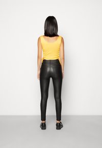 Mladá žena stojí čelem k bílé zdi a má na sobě bezrukávový žlutý top, černé kožené kalhoty s vysokým pasem a černé boty.