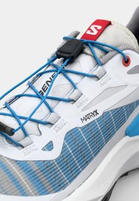 Chaussure de sport avec un dessus en maille bleu et gris, des lacets ronds, un clip de fixation noir et une semelle intermédiaire blanche avec un motif texturé.