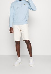 Sudadera con capucha de color azul claro con bolsillo delantero, combinada con pantalones cortos color crema y zapatillas blancas. Presenta un pequeño logo en el pecho.