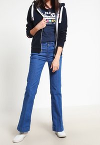 Femme portant un sweat à capuche zippé marine, un t-shirt graphique marine, un jean évasé bleu et des chaussures slip-on blanches, se tenant contre un fond blanc uni.