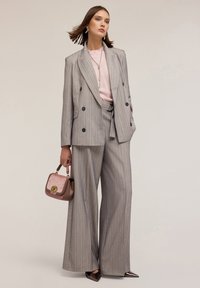 Abito grigio a righe sottili con pantaloni larghi e giacca doppiopetto, abbinato a un maglione rosa chiaro e una piccola borsa bicolore.