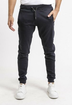 Homme portant un pantalon de jogging noir avec taille à cordon et des baskets blanches, debout avec une main dans la poche et l'autre détendue.