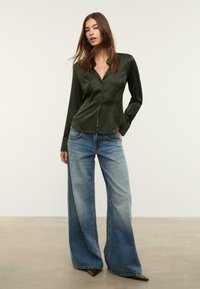 Camicia satinata verde scuro a maniche lunghe con bottoni frontali, abbinata a jeans in denim blu a gamba larga e tacchi a punta neri su uno sfondo neutro.