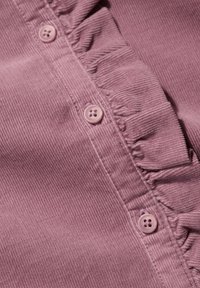 Rožinė kordūrinė medžiaga turi raukšlėtą kraštą palei mygtukų plokštelę, su šviesiai rožiniais mygtukais ir vertikaliu raukšlėtu tekstūra.