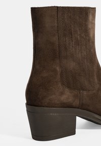 Botte de cheville en cuir suédé marron avec un bout arrondi, un talon bas empilé et des détails de couture verticaux à l'arrière. Semelle en caoutchouc lisse.