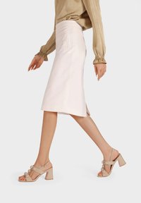 Sandali con tacco alto beige a strappo, con dettagli circolari, tacco a blocco quadrato e finitura lucida. Indossati con una gonna di colore neutro.