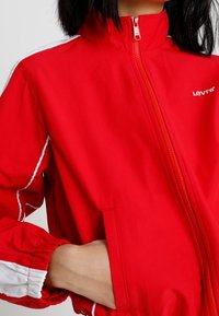 Czerwona kurtka na zamek wykonana z lekkiego materiału, z białymi akcentami, wysokim kołnierzem i elastycznymi mankietami oraz widocznymi kieszeniami.