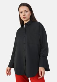 Camisa negra de botones con un bolsillo en el pecho, mangas largas y un corte holgado, hecha de tela suave. Llevada con pantalones rojos.