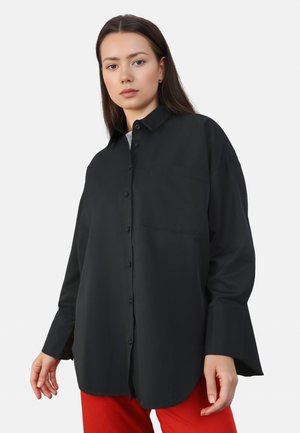Black button-up shirt with a chest pocket, long sleeves, and a loose fit, made from smooth fabric. Worn with red pants.
