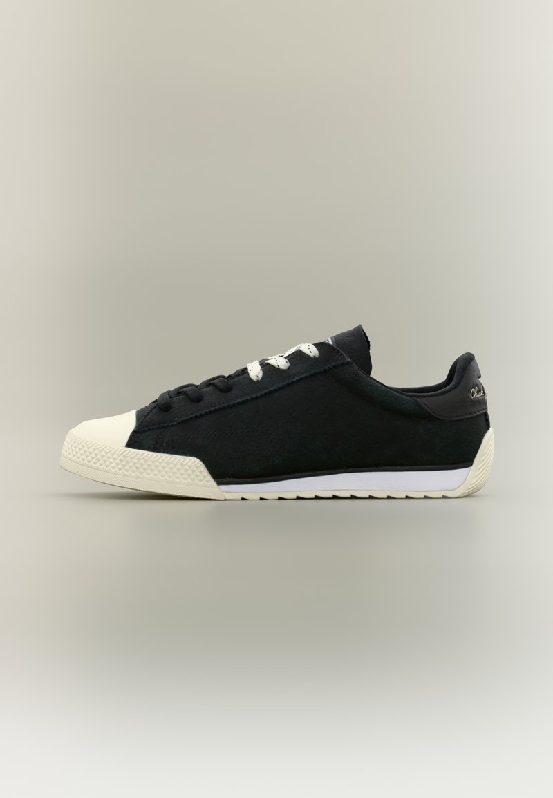 Μαύρο αθλητικό παπούτσι χαμηλού τύπου με λευκή μύτη, ασύμμετρα μαύρα και λευκά κορδόνια, λευκή σόλα και διακριτικό λογότυπο στη φτέρνα.